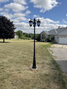 Black decorative three-head lamp post in suburban front yard near driveway and house under blue sky with clouds
