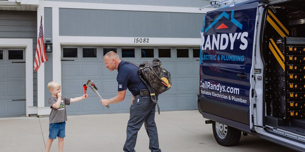 A plumber interacts with a child holding a wrench beside a Randy's Electric & Plumbing van in a suburban driveway.