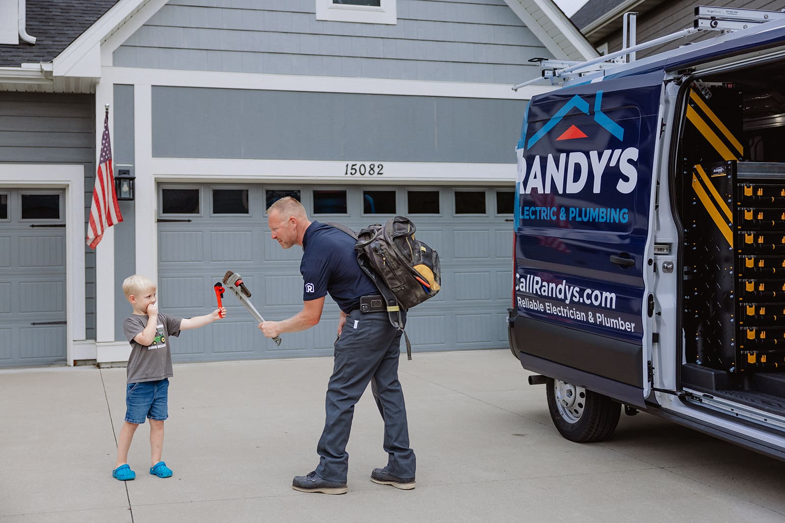 A plumber interacts with a child holding a wrench beside a Randy's Electric & Plumbing van in a suburban driveway.