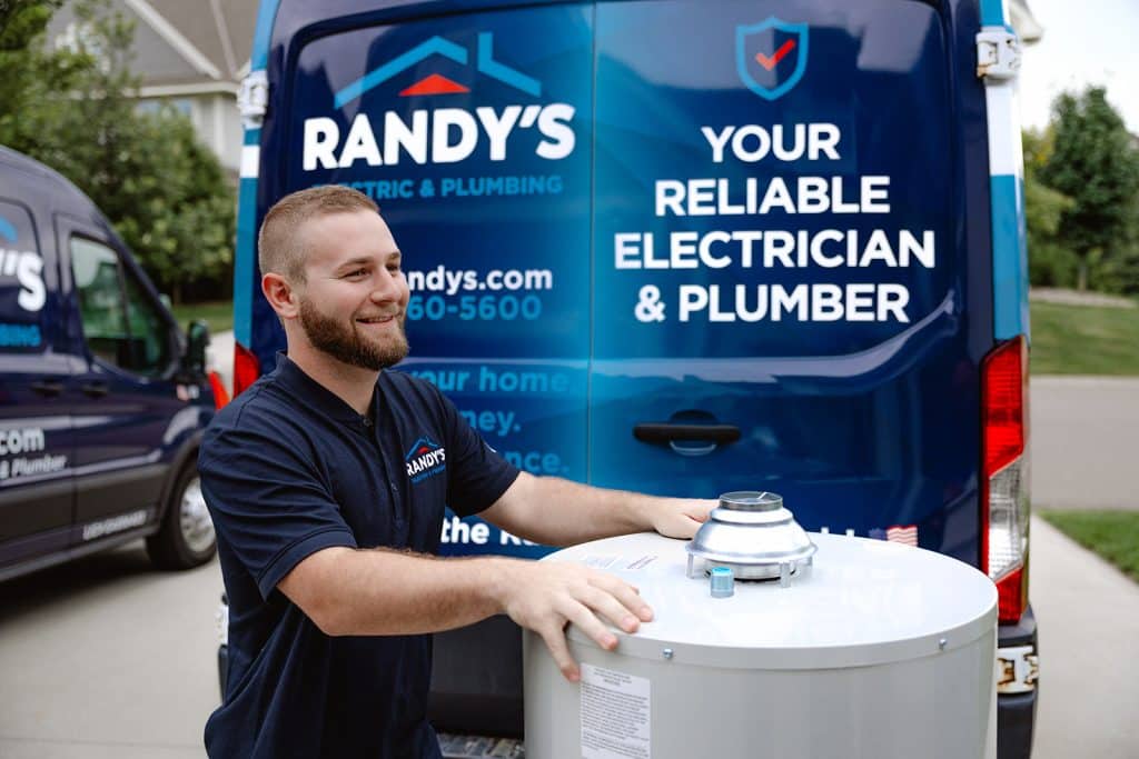 Smiling technician installs water heater beside Randy’s Electric & Plumbing service van in a residential driveway