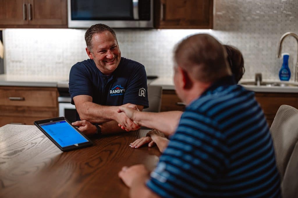 Home services contractor shakes hands with a couple at a kitchen table, tablet open during consultation.