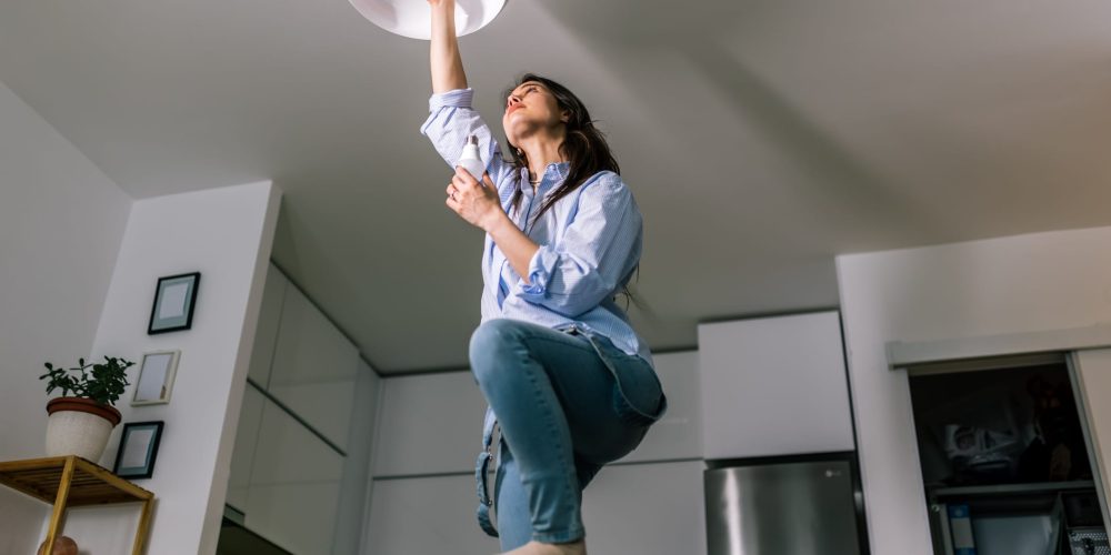 A woman on a ladder checking her lightbulbs to see why they are dimming when her appliances turn on.