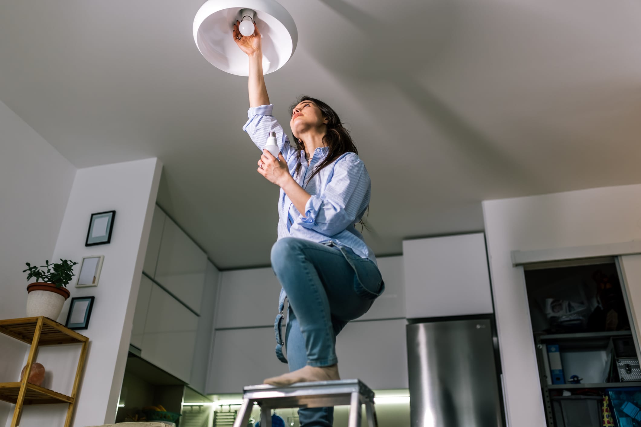 A woman on a ladder checking her lightbulbs to see why they are dimming when her appliances turn on.